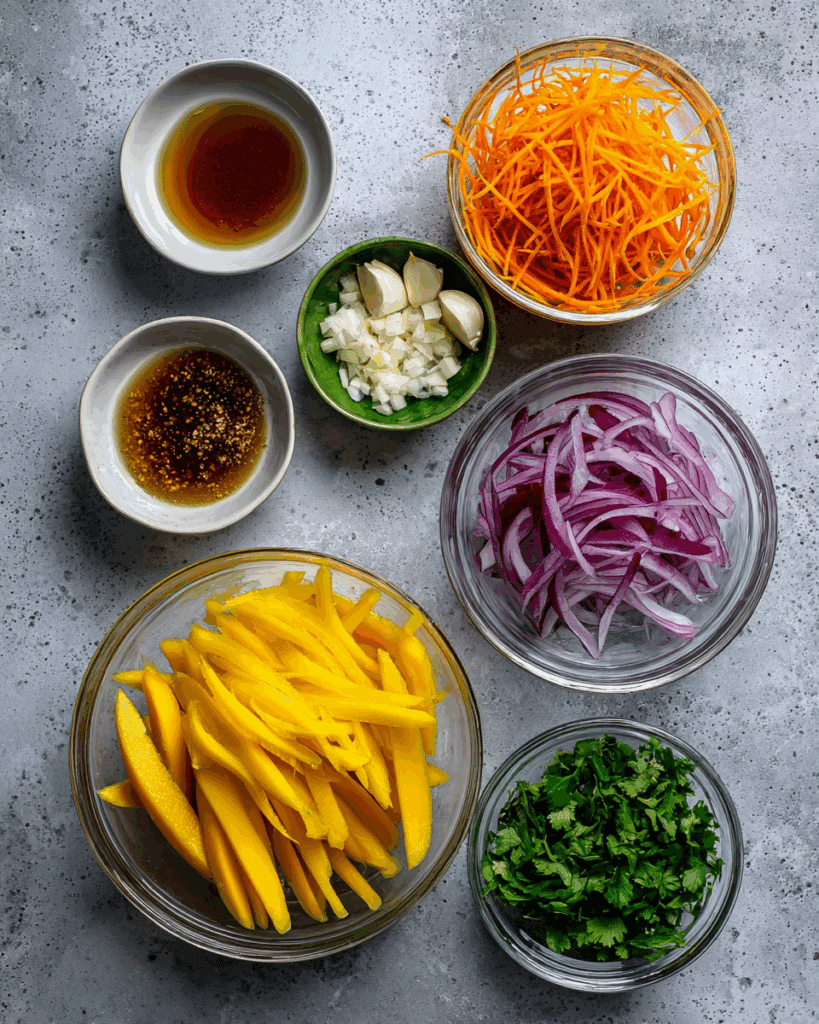 Fresh ingredients for Green Thai Mango Salad including green mangoes, carrots, red onion, lime, chilies, garlic, cilantro, and fish sauce